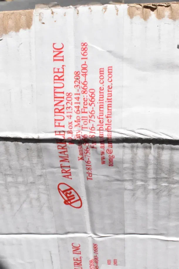 Cardboard box with "Art Marble Furniture, Inc." label in red text and contact information. Box shows signs of wear.