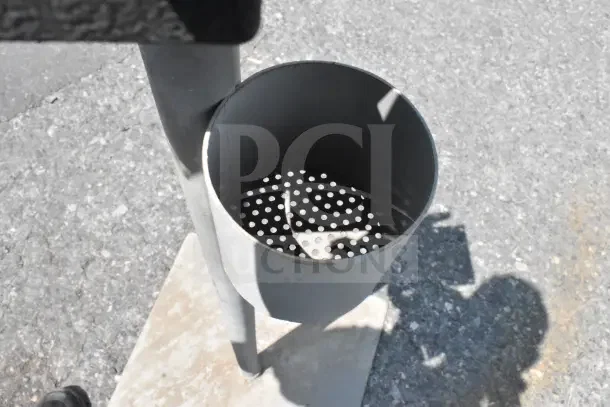 Metal stand with cylindrical holder on concrete base, used condition. Features perforated insert for drainage or support.