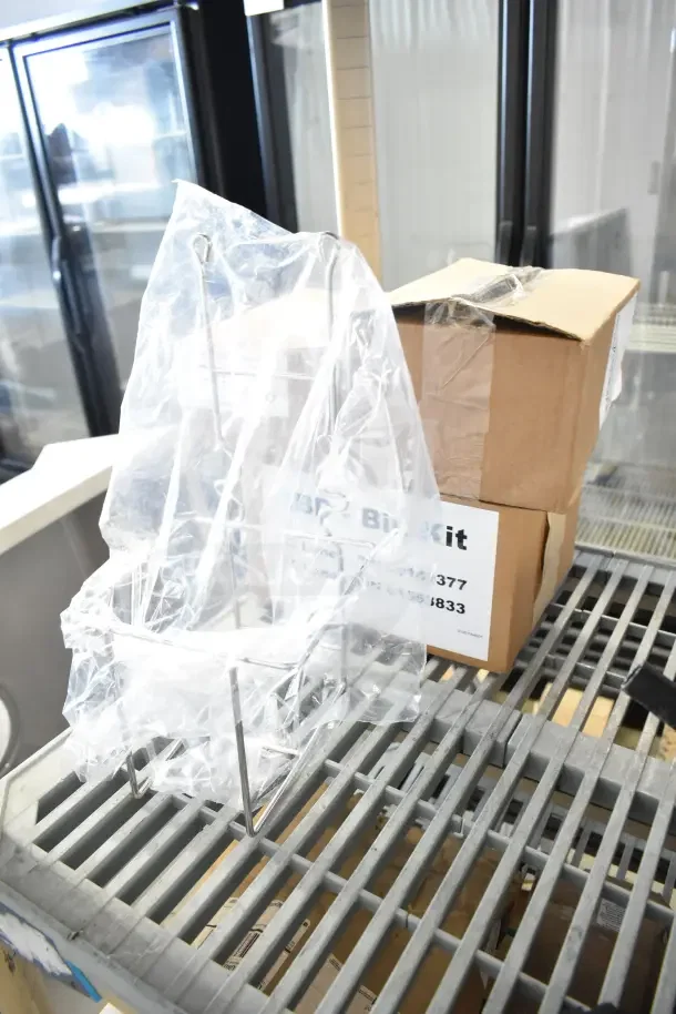 Four brand new boxed items, including a soap dispenser, metal rack, and IBD Bin Kit, shown on a metal grate surface.