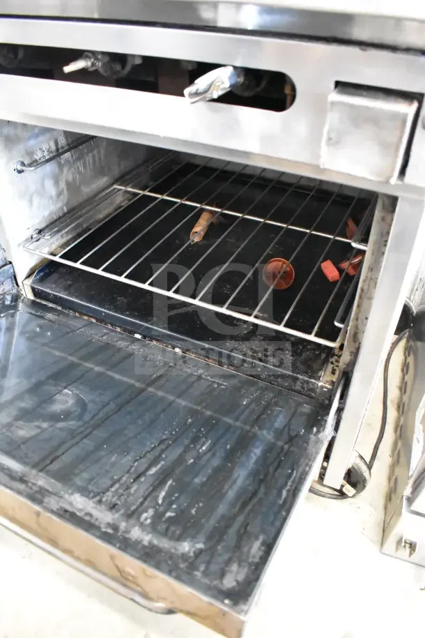 Commercial gas-powered range with 4 burners, flat top griddle, 2 ovens, and backsplash. Shows interior of oven, fair condition.