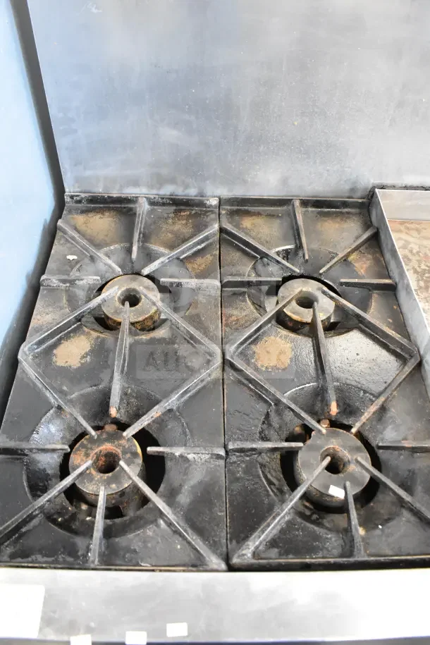Commercial gas-powered range with 4 burners, flat top griddle, 2 ovens, and backsplash. Used condition with visible wear.
