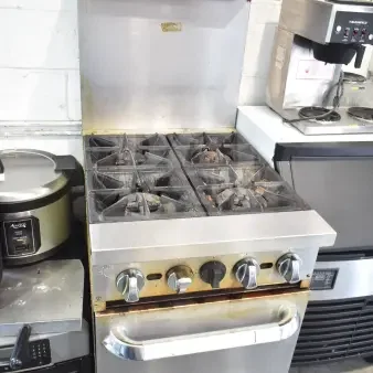 Commercial gas-powered 4 burner range with oven, over shelf, and backsplash. Sturdy, used condition with visible wear.