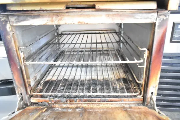 Commercial gas-powered 4 burner range with oven, over shelf, and backsplash, showing interior racks and used condition.