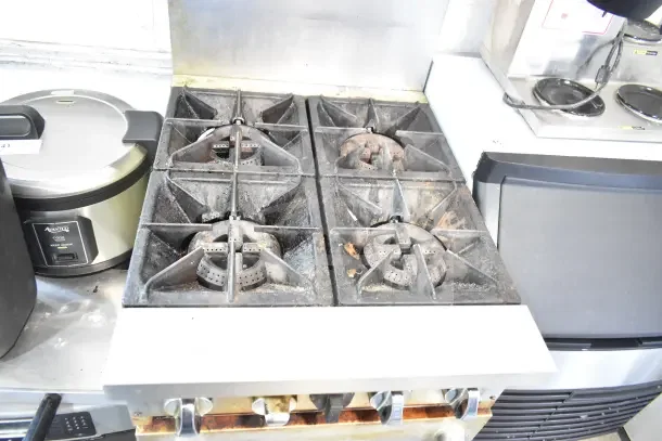 Commercial gas-powered 4-burner range with oven, over-shelf, and backsplash. Visible wear, ideal for busy kitchens.