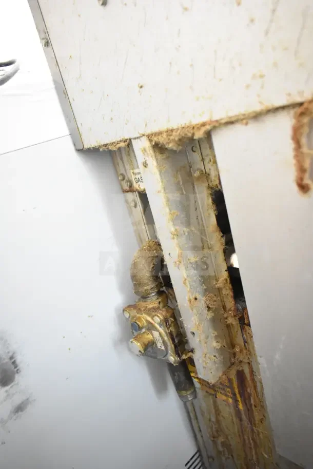 Commercial 4 burner gas range with oven, over shelf, and backsplash. Visible wear and rust on the side panel and connections.