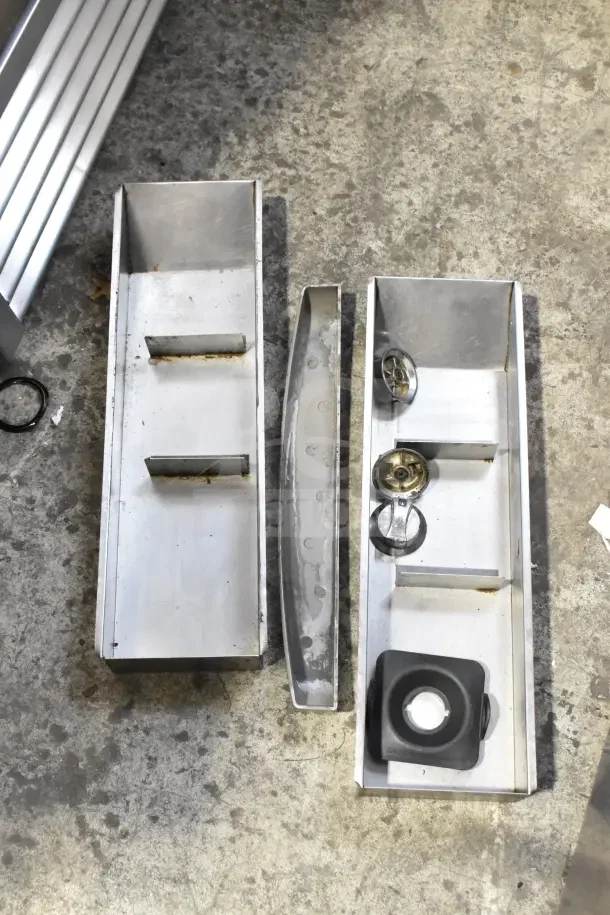 Metal rectangular trays with wheels and parts laid out on concrete floor, part of 25-item kitchenware auction lot.