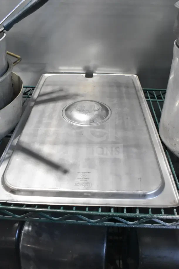 Stainless steel kitchenware assortment on a shelf, featuring a large stock pot, lid with handle, and visible Holikme branding.