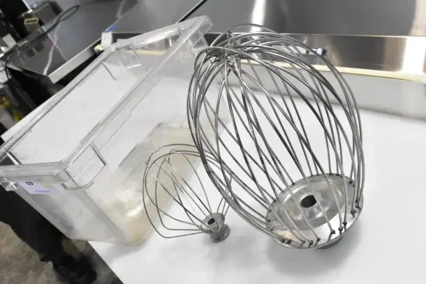 Mixer whisk attachments in steel, varying sizes, next to clear storage bin, part of 25-item auction lot.