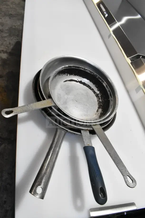 Stack of skillets with metal and plastic handles, showing visible wear, part of a 25-item auction lot.